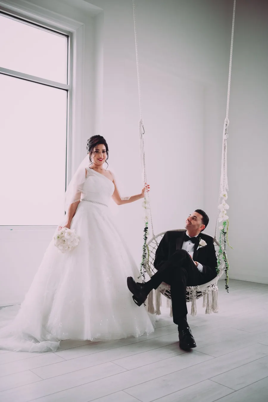 Bride standing beside groom seated on a macrame swing in a bright white studio — creative wedding portrait photography Niagara by Captivate Photo+Cinema