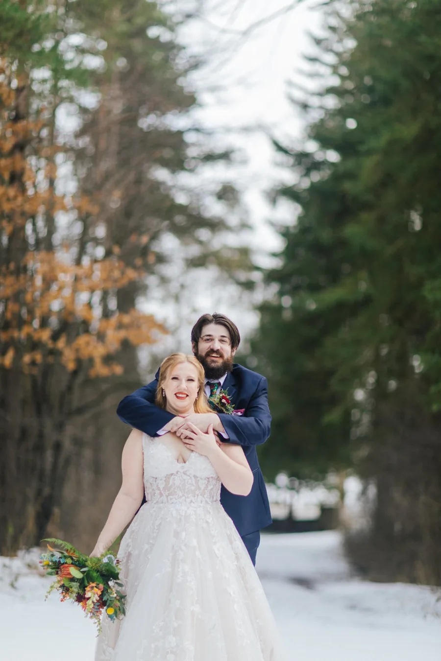 Bride and groom embracing on a snowy lane surrounded by autumn and pine trees — winter wedding photography Niagara Region by Captivate Photo+Cinema